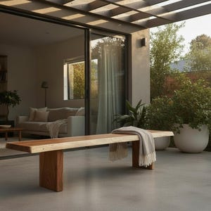May include: A wooden bench with a natural finish and a rectangular top. The bench has a dark brown base and a light brown top. A light gray blanket with fringe is draped over the bench. The bench is outdoors on a concrete patio.
