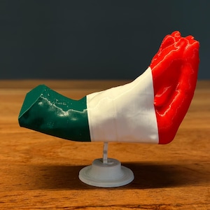 Op de afbeelding: Een 3D-geprinte sculptuur van de Italiaanse vlag, met groene, witte en rode secties. De vlag is gemonteerd op een kleine, witte standaard en wordt weergegeven op een houten oppervlak.