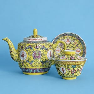 May include: A yellow ceramic teapot and a matching lidded bowl with a saucer, featuring intricate floral patterns in pink, green, and blue, along with red Chinese characters. The set displays a repeating geometric border design in black and white.