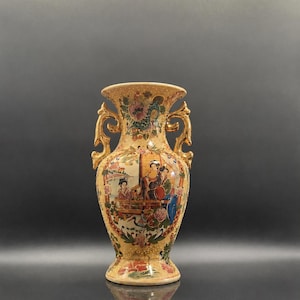 Puede incluir: Un jarrón de cerámica con base de color beige, que presenta dos asas de color dorado y un borde acampanado. La superficie está decorada con coloridas ilustraciones de figuras con vestimenta tradicional, motivos florales y puntos dorados en relieve.