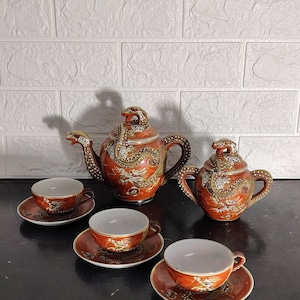 Könnte beinhalten: Ein Teeservice bestehend aus einer Teekanne, einer Zuckerdose mit Deckel und drei Teetassen mit passenden Untertassen. Jedes Stück ist aus orangefarbener Keramik gefertigt und mit goldenen und weißen Drachenmotiven sowie Reliefstrukturen verziert.