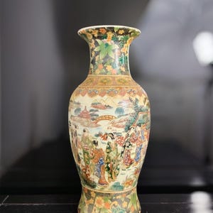 Puede incluir: Un jarrón de cerámica con una escena policromada detallada de figuras con vestimenta tradicional en un paisaje. El cuello y la base están decorados con densos patrones florales en negro, verde y naranja. El cuerpo central representa un puente, edificios y árboles.