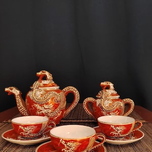 Op de afbeelding: Een theeservies bestaande uit een theepot, een suikerpot met deksel en drie theekopjes met bijpassende schoteltjes. Elk stuk is afgewerkt in een dieporanje glazuur met gedetailleerde gouden en witte drakenmotieven, gouden randen en draakvormige dekselknoppen.