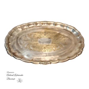 Pode incluir: Uma bandeja oval prateada com um padrão floral decorativo no centro. A bandeja tem uma borda ondulada e uma aparência ligeiramente manchada. A bandeja é provavelmente vintage ou antiga.