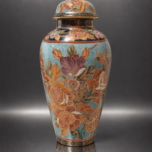 May include: A decorative ceramic ginger jar with a lid, featuring a light blue crackle glaze background adorned with detailed floral patterns in shades of orange, purple, and gold, finished with dark blue and gold geometric borders at the rim and base.