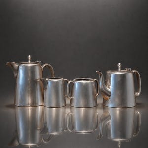 Op de afbeelding: Een vierdelige zilverkleurige thee- en koffieset bestaande uit een hoge koffiepot, een kleinere theepot, een melkkan en een suikerpot. Elk item heeft een glad, cilindervormig lichaam, een gebogen handvat en een deksel met een knop.