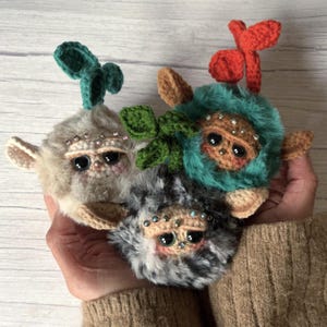 May include: Three handcrafted plush toys with large black eyes, crocheted faces, and faux fur bodies. Each has a different colour scheme: grey, teal, and a mix of grey and white. They are decorated with small embellishments and crocheted leaf accents.