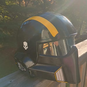 Peut inclure: Casque noir avec une bande jaune et un emblème de crâne. Le casque a une visière et une mentonnière. Le casque est posé sur une surface en bois. Le casque a un design futuriste.