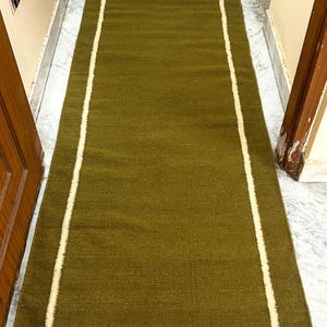 Puede incluir: Alfombra rectangular verde oliva con un borde crema a cada lado. La alfombra está hecha de un material tejido y está colocada sobre un suelo de mármol. Es una alfombra larga y estrecha.