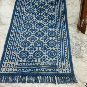 May include: A blue and white patterned runner rug with a floral design. The rug has a fringe on the bottom edge and a border design. The rug is made of a woven material and is rectangular in shape.