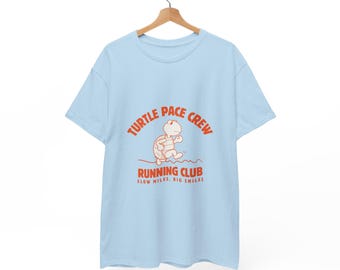 May include: Light blue t-shirt with the text "TURTLE PACE CREW RUNNING CLUB" in orange. The shirt features a cartoon turtle graphic. The text below the graphic reads "SLOW MILES, BIG SMILES". The shirt is hanging on a wooden hanger.