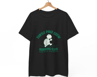 May include: Black t-shirt with a green graphic of a running turtle and the text "Turtle Pace Crew Running Club Slow Miles, Big Smiles". The shirt is hanging on a wooden hanger.