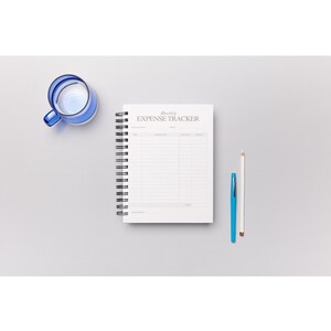 May include: A spiral-bound notebook with the words "Monthly Expense Tracker" printed on the cover. A blue pen, a white pencil, and a blue glass with liquid are next to the notebook on a grey surface.