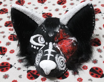 Könnte beinhalten: Eine Tiermaske aus schwarzem Kunstfell mit großen spitzen Ohren und weiß strukturierten Rändern. Das Gesicht zeigt weiß gemalte Spiral- und Linienmuster, eine weiße Nase und einen auffälligen roten, strukturierten Fleck am linken Auge.