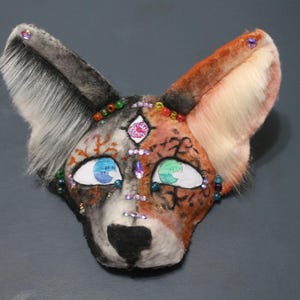 May include: A handcrafted, decorative mask in the shape of a fox's face. The mask features a color scheme of black, gray, orange, and white, with detailed eyes and embellished with colorful beads and gems. The ears are fluffy.