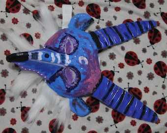 Könnte beinhalten: Eine handgefertigte Maske mit einer blau und violett bemalten Oberfläche, einer langen Schnauze, zwei großen, geschwungenen Hörnern mit schwarzen Streifen und weißen Kunstfellakzenten. Die Maske zeigt dunkle Augenhöhlen und eine spitze Nase, was ein kreaturenartiges Aussehen mit strukturierter Oberfläche und lebendiger Farbgebung erzeugt.