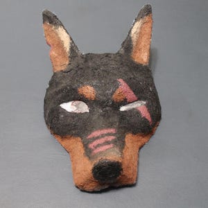 May include: A handmade dog mask with a black and brown color scheme. The mask features a detailed design with visible ears, eyes, and a snout. The dog's face has red markings and a textured surface, suggesting it's made from a natural material.