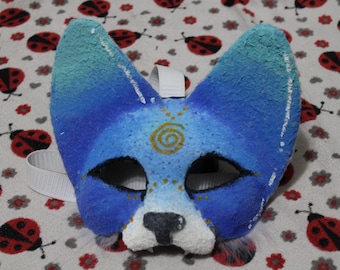Könnte beinhalten: Eine handgefertigte Tiermaske mit strukturierter Oberfläche in einem Blau- und Türkisverlauf. Die Maske zeigt ein goldenes Spiralsymbol auf der Stirn, schwarze Augenumrandungen und eine weiße Schnauze mit schwarzer Nase. Inklusive weißem Gummiband.