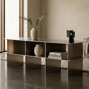 May include: A modern, metallic coffee table with open cubby storage. The table holds two ceramic vases, a small sculpture, and a stack of books. The room has a neutral color palette with a rug and a window.