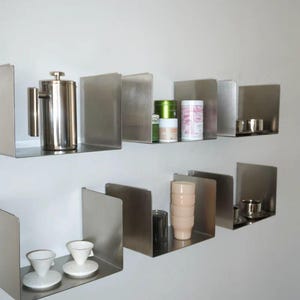 May include: Stainless steel floating shelves with various kitchen items. A French press, tea canisters, cups, and saucers are displayed. The shelves are mounted on a white wall, creating a modern, minimalist aesthetic.