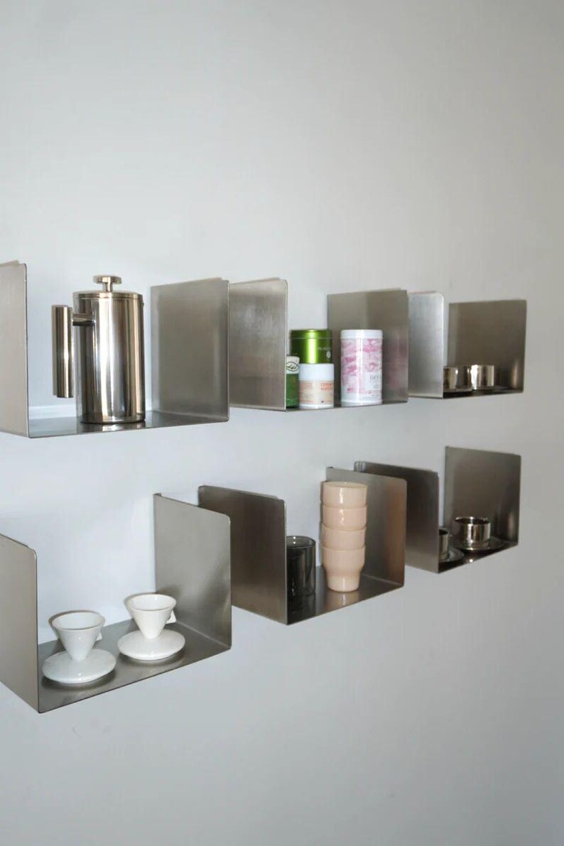 May include: Stainless steel floating shelves with various kitchen items. A French press, tea canisters, cups, and saucers are displayed. The shelves are mounted on a white wall, creating a modern, minimalist aesthetic.