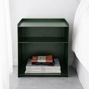 May include: A dark forest green metal nightstand featuring a rectangular frame with a raised edge on the top surface and a single horizontal shelf. The lower section contains a stack of various books and magazines with visible text on the spines.