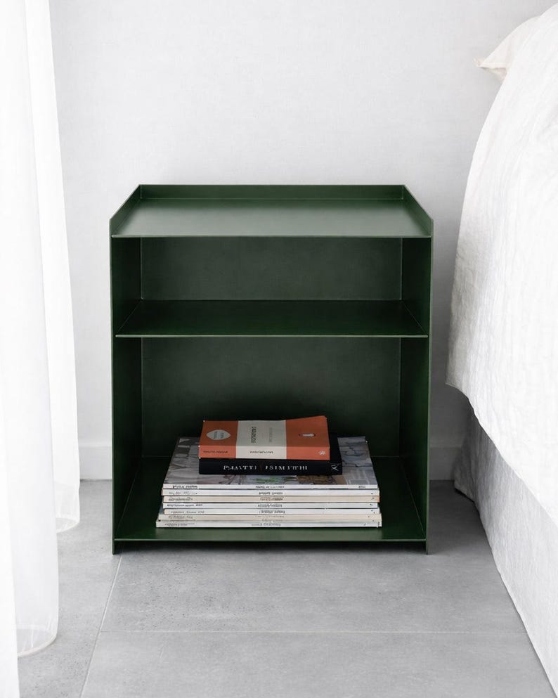 Puede incluir: Una mesita de noche de metal en color verde bosque oscuro con un marco rectangular, un borde elevado en la superficie superior y un estante horizontal. La secci&oacute;n inferior contiene una pila de varios libros y revistas con texto visible.
