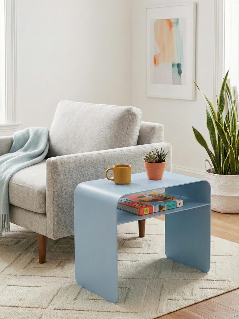 May include: A light blue, modern side table with a curved design. A yellow mug and a small potted succulent sit on top. A colorful book is on the lower shelf. The table is in a living room setting with a gray armchair and a patterned rug.