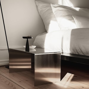 May include: A rectangular side table constructed from a single piece of brushed stainless steel with a bent design. The table features a smooth, reflective metallic surface and sits low to the ground, serving as a bedside stand next to a bed.