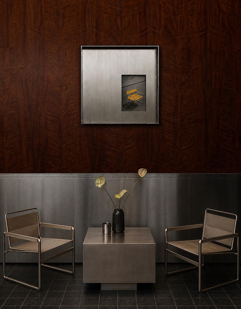 May include: Two metal chairs with light-colored fabric seats and backrests flank a square metal side table. On the table sits a dark vase with three yellow flowers and a small metal cup. A square metal-framed artwork hangs on the wall above.