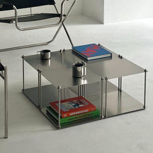 Op de afbeelding: Een moderne vierkante salontafel van roestvrij staal met twee niveaus. De bovenste laag bevat twee zilveren mokken en een blauw boek met rode tekst. De onderste laag toont een stapel boeken met kleurrijke covers. De tafel wordt ondersteund door dunne, cilindrische poten.