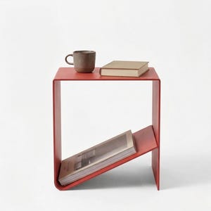 Può includere: Un tavolino rosso moderno con un portariviste integrato. Una tazza marrone e un libro sono sul piano del tavolo. Il tavolo è in metallo e ha un design minimalista. Il tavolo è alto circa 50 cm.