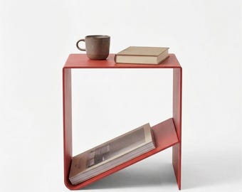 May include: A modern, red side table with a built-in magazine rack. A brown mug and a book rest on the tabletop. The table is made of a single piece of metal and has a minimalist design.