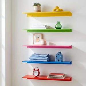 Op de afbeelding: Een reeks van vijf felgekleurde wandplanken in geel, groen, roze, blauw en rood. Elke plank bevat decoratieve items zoals een vetplant, citroenen, boeken, een vaas, een ingelijste print, een kaars, gevouwen linnengoed, een keramisch huis, een wekker en notitieboekjes.