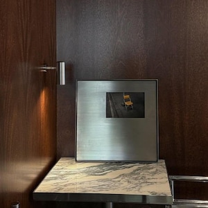 May include: A square, brushed metal frame containing a photograph of a yellow chair on a dark surface. The frame is positioned on a square table with a white and grey marble top, set against a dark wood paneled wall with a small silver wall lamp.