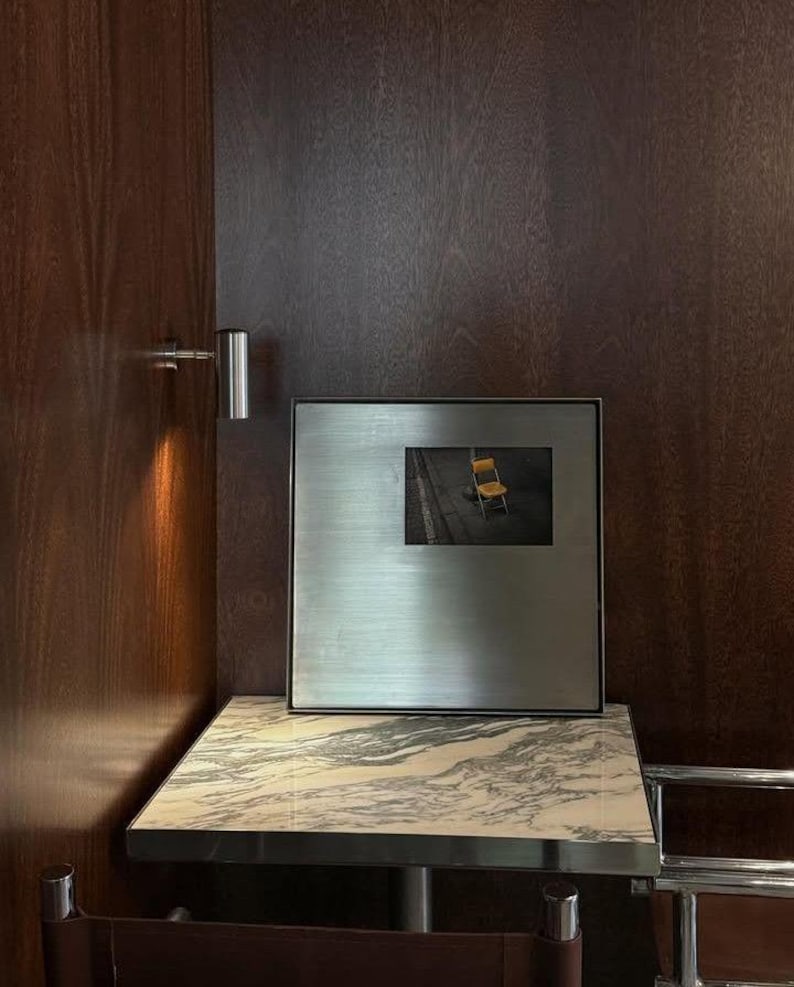 May include: A square, brushed metal frame containing a photograph of a yellow chair on a dark surface. The frame is positioned on a square table with a white and grey marble top, set against a dark wood paneled wall with a small silver wall lamp.