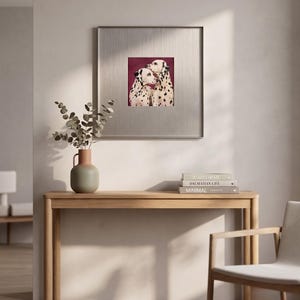 May include: A square framed wall art piece featuring a painting of two Dalmatian dogs with black spots against a deep burgundy background. The artwork is encased in a metallic silver frame with a wide mat border, displayed on a light wall.