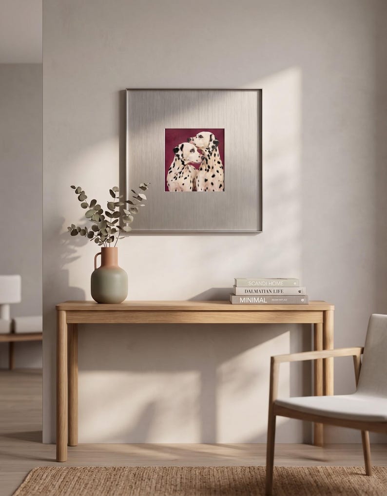 May include: A square framed wall art piece featuring a painting of two Dalmatian dogs with black spots against a deep burgundy background. The artwork is encased in a metallic silver frame with a wide mat border, displayed on a light wall.