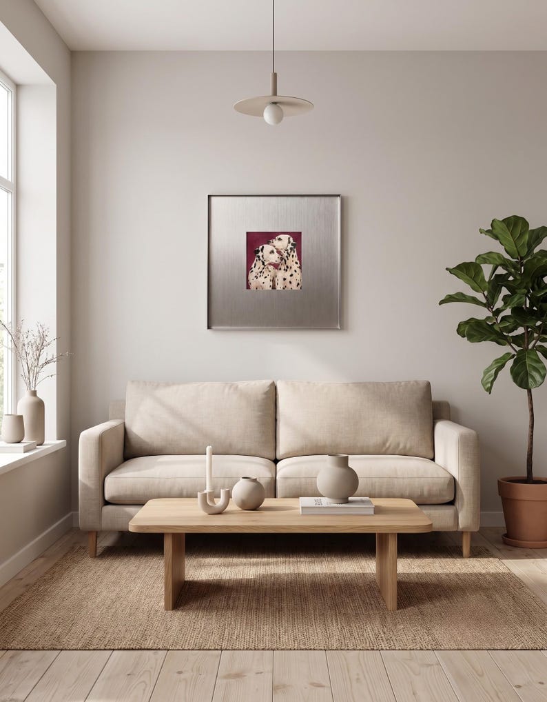May include: A square framed wall art piece featuring two Dalmatian dogs against a deep red background, displayed above a beige sofa, a light wood coffee table with a candle and vase, and a potted plant in a bright, neutral-toned room.