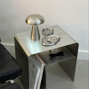 Può includere: Un tavolino moderno in metallo spazzolato con una lampada a forma di fungo, un posacenere e un vassoio decorativo con gioielli. Il tavolo ha anche un ripiano inferiore con due libri e occhiali da sole.
