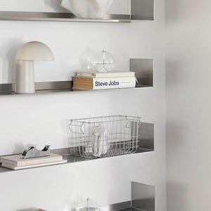 May include: A modern wall display featuring four stainless steel shelves. The shelves hold various decorative items, including a white sculpture, a mushroom-shaped lamp, a book titled "Steve Jobs", a wire basket, and a small square container.