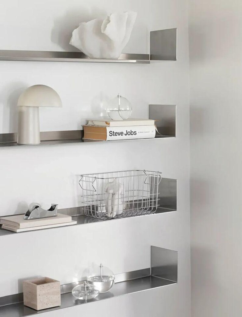 May include: A modern wall display featuring four stainless steel shelves. The shelves hold various decorative items, including a white sculpture, a mushroom-shaped lamp, a book titled "Steve Jobs", a wire basket, and a small square container.