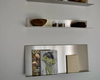 May include: Three modern, stainless steel floating shelves mounted on a white wall. The top shelf holds a dark mug and small wooden objects. The middle shelf has a wooden bowl and metal cylinders. The bottom shelf displays magazines and a book.