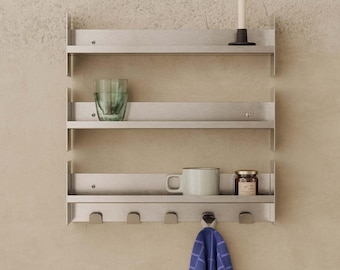 May include: A three-tiered brushed metal wall shelf featuring a top shelf with a white candle, a middle shelf with a green glass tumbler, and a bottom shelf with a white mug, a small glass jar, and five hooks, one holding a blue checkered cloth.