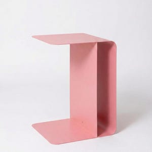 May include: A modern, minimalist side table in a soft pink hue. The table has a C-shaped design with a flat top, a vertical support, and a base. The table measures approximately 20 inches long, 12 inches wide, and 24 inches high.