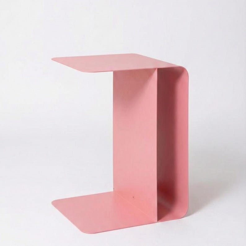 May include: A modern, minimalist side table in a soft pink hue. The table has a C-shaped design with a flat top, a vertical support, and a base. The table measures approximately 20 inches long, 12 inches wide, and 24 inches high.