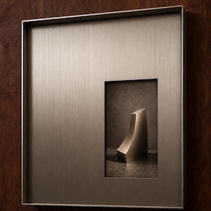 May include: A square, brushed metallic wall frame with a vertical rectangular cutout. Inside the cutout, a small, curved, solid metallic wedge-shaped object sits on a dark, textured surface, creating a dimensional display piece.