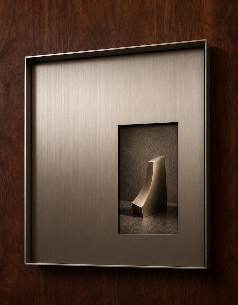 May include: A square, brushed metallic wall frame with a vertical rectangular cutout. Inside the cutout, a small, curved, solid metallic wedge-shaped object sits on a dark, textured surface, creating a dimensional display piece.