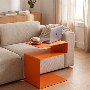 Puede incluir: Una mesa auxiliar naranja con un diseño de dos niveles. Se coloca junto a un sofá beige, con un portátil y una taza de café en el nivel superior. El diseño moderno y el color brillante la convierten en una pieza de acento elegante.