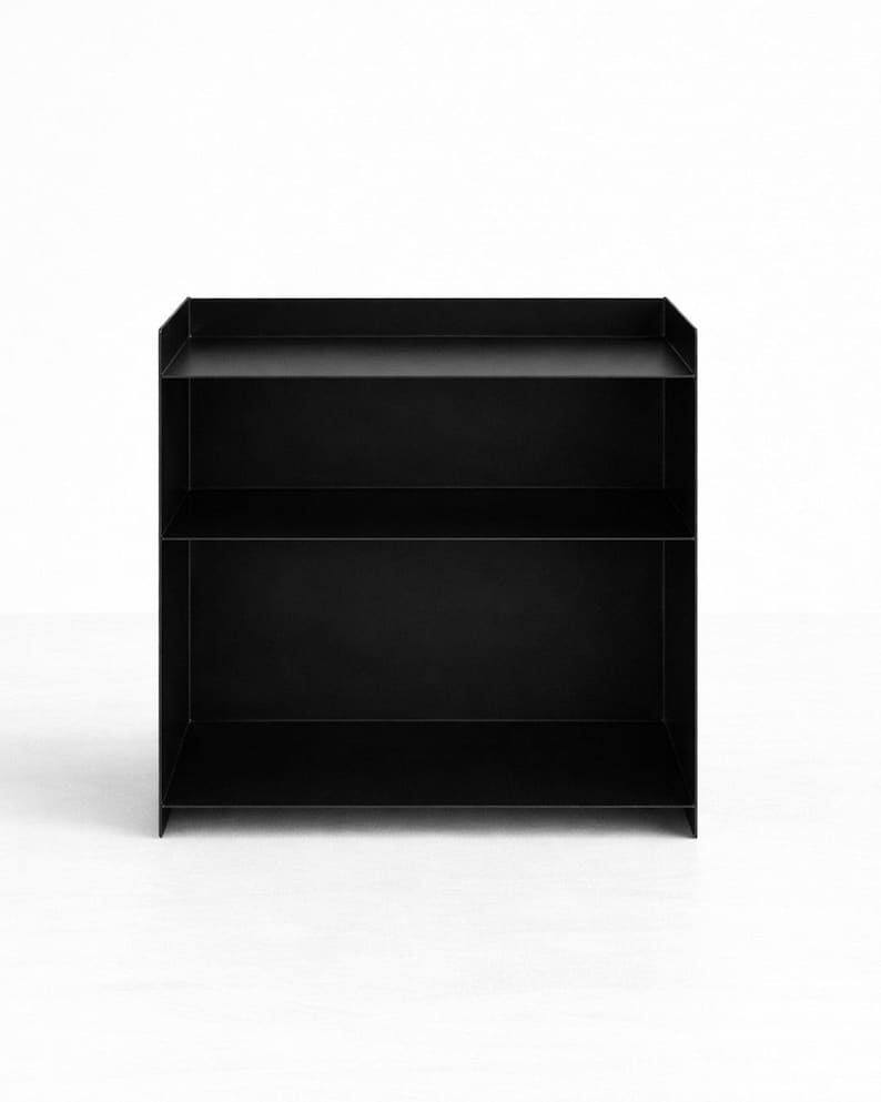 Puede incluir: Una estanter&iacute;a de metal negro mate con dos niveles de almacenamiento abiertos y una superficie superior con bordes laterales elevados. La estructura est&aacute; compuesta por paneles finos y planos con un marco rectangular y un acabado oscuro uniforme.